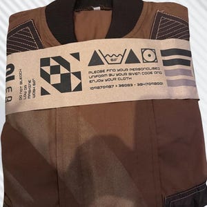 May include: A brown jacket with a dark brown ribbed collar and geometric stitching details on the shoulders. A tan paper band wraps around the center, featuring black graphic icons, care instructions, and the text: PLEASE FIND YOUR PERSONALISED UNIFORM BY YOUR GIVEN CODE AND ENJOY YOUR CLOTH 109870987 - 36083 - 3847098001.