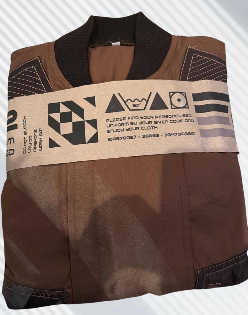 May include: A brown jacket with a dark brown ribbed collar and geometric stitching details on the shoulders. A tan paper band wraps around the center, featuring black graphic icons, care instructions, and the text: PLEASE FIND YOUR PERSONALISED UNIFORM BY YOUR GIVEN CODE AND ENJOY YOUR CLOTH 109870987 - 36083 - 3847098001.