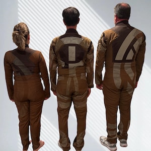 May include: Three individuals seen from the back wearing matching brown jumpsuits with long sleeves and high collars. The jumpsuits feature large, geometric, light tan and dark brown fabric panels and patches arranged in abstract patterns across the back.