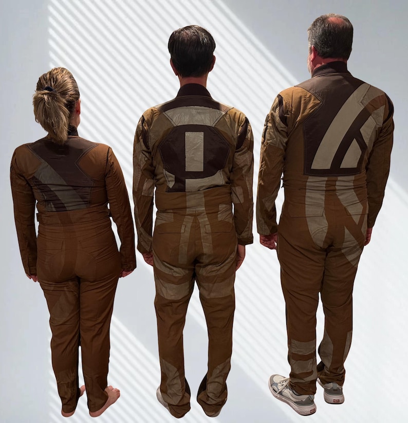 May include: Three individuals seen from the back wearing matching brown jumpsuits with long sleeves and high collars. The jumpsuits feature large, geometric, light tan and dark brown fabric panels and patches arranged in abstract patterns across the back.