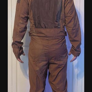 May include: A brown jumpsuit with a dark brown back panel and long sleeves, paired with a weathered green and silver helmet featuring a T-shaped visor and a side-mounted antenna. The outfit is worn with grey and black hiking boots.