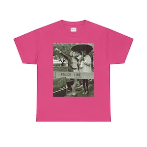 Puede incluir: Una camiseta rosa intenso con una imagen en blanco y negro de dos personas detrás de una línea policial. La imagen muestra el texto "POLICE LINE" en una barrera. La camiseta tiene cuello redondo clásico y mangas cortas.