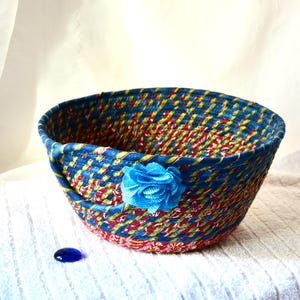 May include: A handmade, woven basket with a vibrant mix of blue, red, and yellow threads. A blue fabric flower adorns the side. The basket sits on a white surface, with a blue glass gem nearby. This decorative basket is ideal for storage or display.