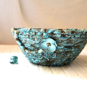 May include: A round bowl constructed from coiled fabric strips featuring a teal, brown, and white paisley-inspired pattern. A single light blue fabric flower with a small gold-toned center is attached to the exterior side of the bowl.