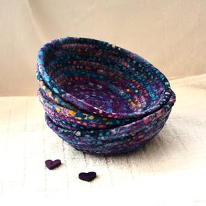May include: A stack of three small, round fabric bowls in shades of purple, blue, and yellow. The bowls have a textured appearance and are decorated with small, colorful dots. Two heart-shaped buttons are placed below the bowls.