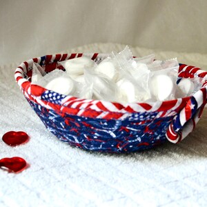 May include: A fabric bowl with a patriotic design, featuring red, white, and blue patterns. The bowl is filled with individually wrapped white candies. Two red heart-shaped decorations are placed near the bowl.
