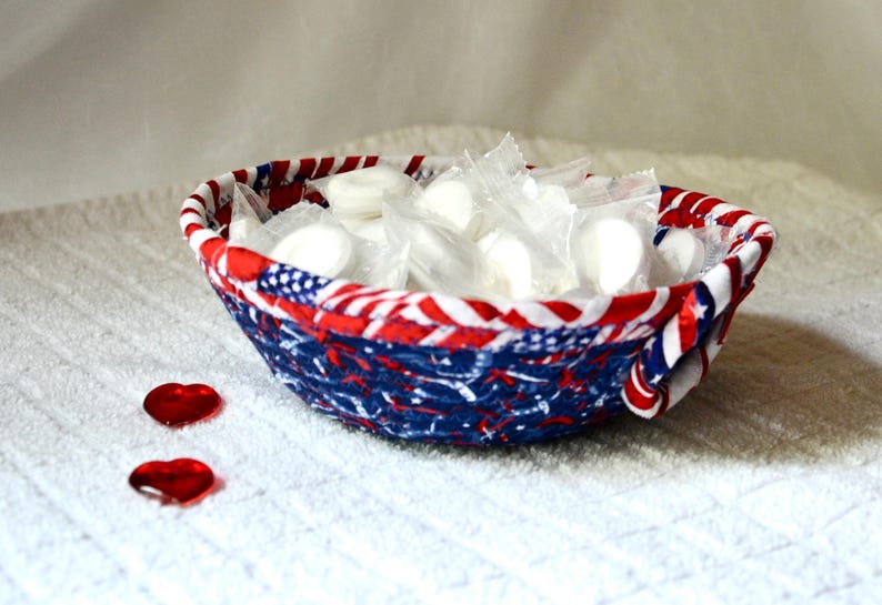 May include: A fabric bowl with a patriotic design, featuring red, white, and blue patterns. The bowl is filled with individually wrapped white candies. Two red heart-shaped decorations are placed near the bowl.