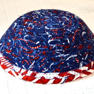 May include: A fabric bowl with a patriotic design. The bowl features a blue background with red and white accents, and a red, white, and blue striped border. The bowl is handmade and has a unique, textured appearance.