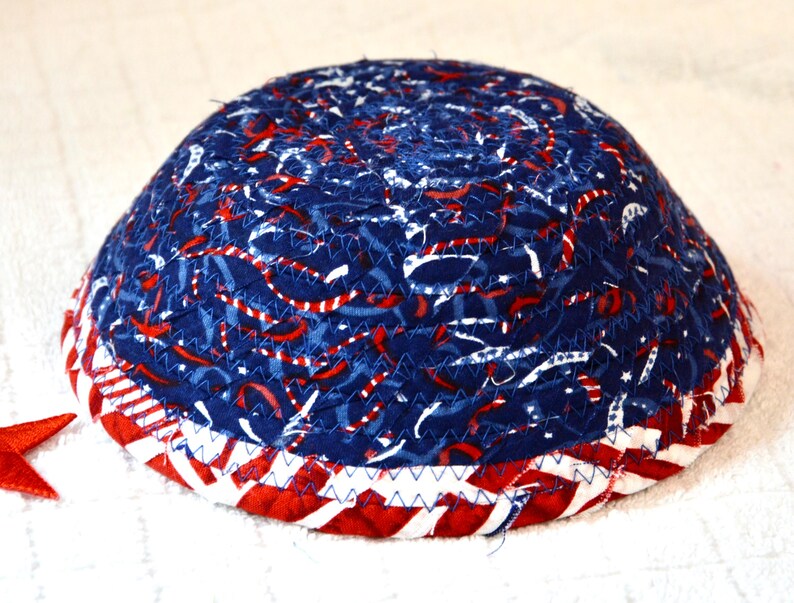 May include: A fabric bowl with a patriotic design. The bowl features a blue background with red and white accents, and a red, white, and blue striped border. The bowl is handmade and has a unique, textured appearance.