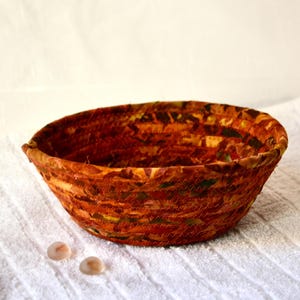 May include: A woven fabric bowl with a brown, orange, and green pattern. The bowl is sitting on a white surface.