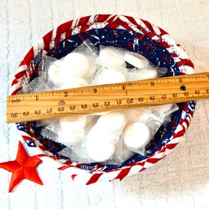 May include: A round, fabric-lined basket with a red, white, and blue patriotic design. The basket contains individually wrapped white items. A wooden ruler, measuring approximately 10 inches, rests inside the basket. A red star is next to the basket.