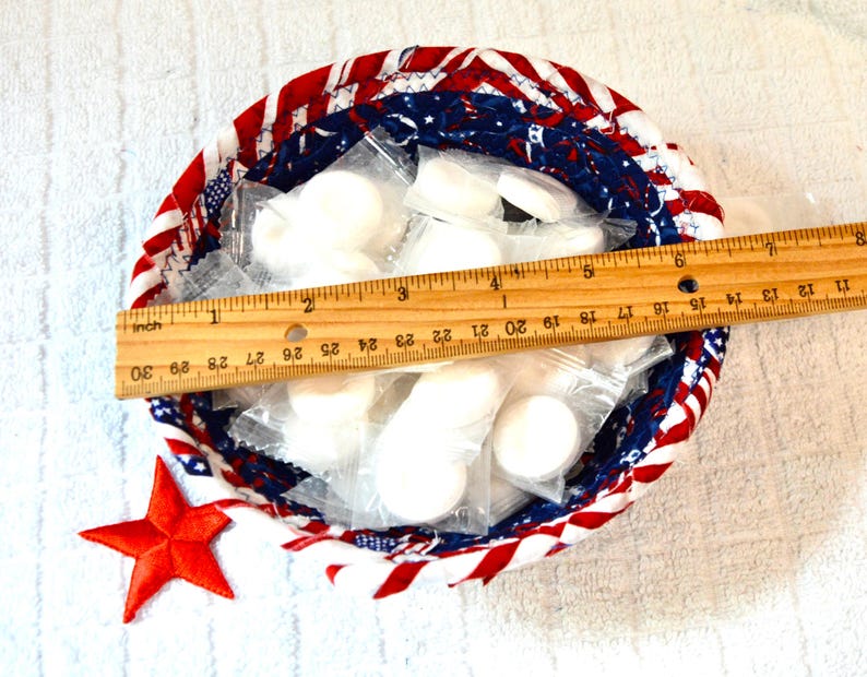 May include: A round, fabric-lined basket with a red, white, and blue patriotic design. The basket contains individually wrapped white items. A wooden ruler, measuring approximately 10 inches, rests inside the basket. A red star is next to the basket.
