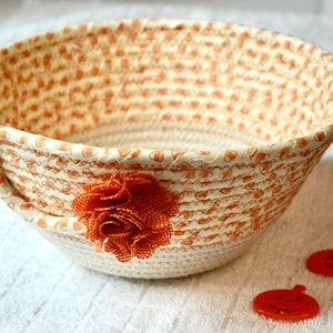 May include: A cream-colored rope bowl with a patterned fabric lining featuring orange candy corn shapes. An orange burlap flower is attached to the side. Two small orange pumpkin-shaped decorations are next to the bowl. A decorative storage basket.