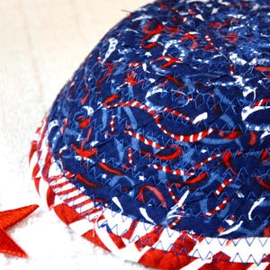 May include: A quilted, hemispherical object with a patriotic theme. The top is navy blue with red and white ribbon-like patterns. The bottom edge features alternating red and white stripes, with blue stitching. A red star is visible.
