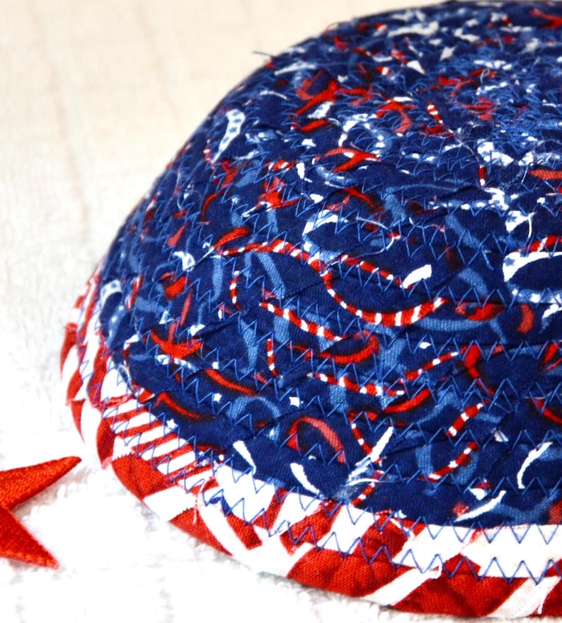 May include: A quilted, hemispherical object with a patriotic theme. The top is navy blue with red and white ribbon-like patterns. The bottom edge features alternating red and white stripes, with blue stitching. A red star is visible.