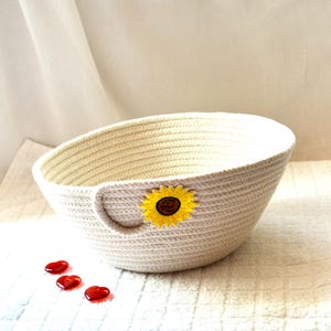 May include: A cream-colored, oval-shaped rope basket with a sunflower applique. The basket has a handle on one side and is decorated with a yellow and brown sunflower. Three red heart-shaped glass beads are in front of the basket.