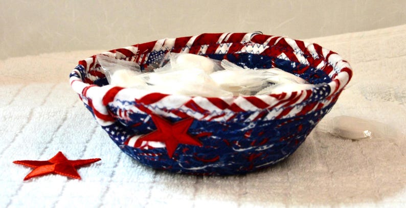 May include: A fabric bowl with a patriotic design, featuring red, white, and blue colors. The bowl is filled with white items and has a red star applique on the side. A separate red star is also visible.