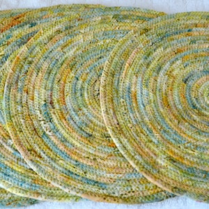 May include: A set of six round placemats. Each placemat is made of a tightly woven, spiraling pattern of fabric in shades of green, yellow, blue, and tan. The placemats are arranged in a row, showcasing their circular design and textured surface.