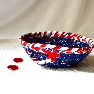 May include: A patriotic, handmade fabric bowl in red, white, and blue. The bowl features a star and stripe design, with a red star prominently displayed. Two red heart-shaped embellishments are near the bowl.