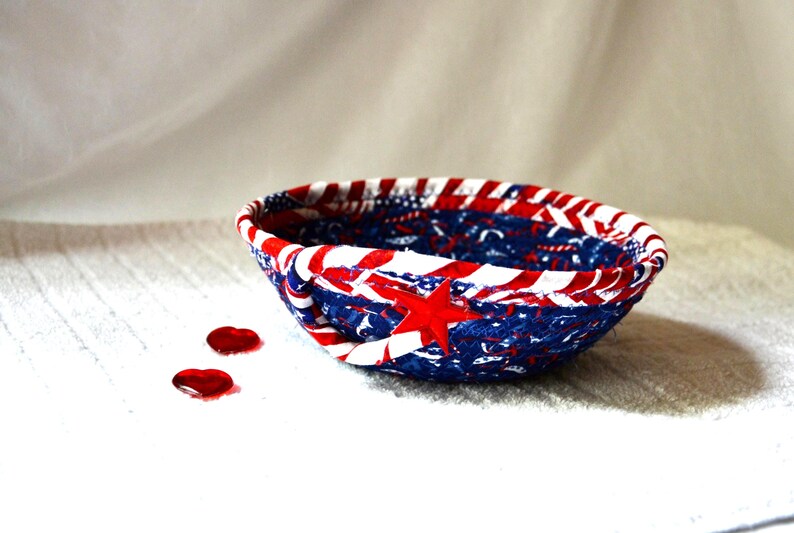 May include: A patriotic, handmade fabric bowl in red, white, and blue. The bowl features a star and stripe design, with a red star prominently displayed. Two red heart-shaped embellishments are near the bowl.
