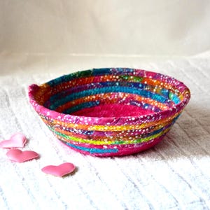 May include: A round, shallow bowl constructed from coiled fabric strips in vibrant shades of pink, orange, yellow, green, and blue, featuring a white dotted pattern throughout. Three small, smooth, pink heart-shaped objects lie nearby.