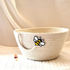 May include: A cream-colored rope basket with a handle and a bee-shaped sticker. The basket is round with a wide opening and sits on a white surface. The bee sticker is black, yellow, and white.