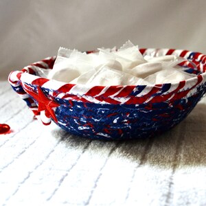 May include: A patriotic-themed bowl with a red, white, and blue design, filled with individually wrapped white candies. A red, star-shaped accent adorns the side. The bowl sits on a white, textured surface.