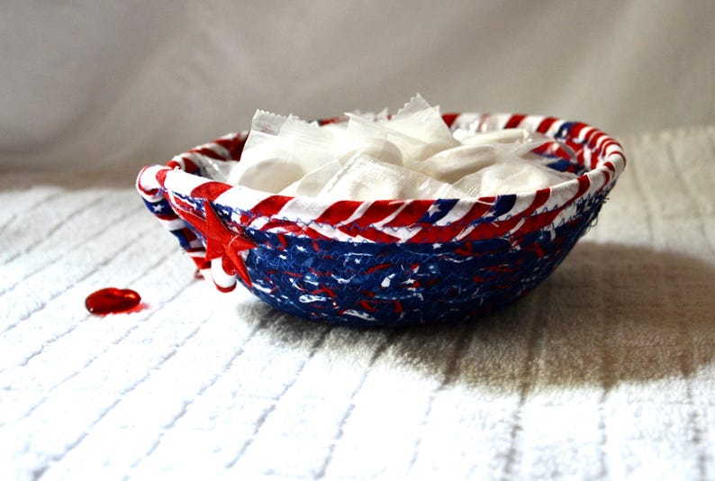 May include: A patriotic-themed bowl with a red, white, and blue design, filled with individually wrapped white candies. A red, star-shaped accent adorns the side. The bowl sits on a white, textured surface.