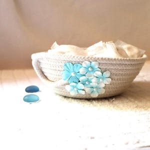 May include: A small, round, off-white woven rope basket decorated with a cluster of light blue and white artificial flowers. The basket contains several small, wrapped white items, and two smooth blue stones are placed beside the basket.