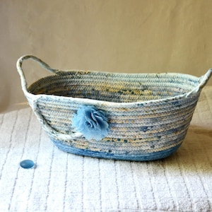May include: An oval-shaped basket constructed from coiled rope with a variegated blue, white, and yellow pattern. The basket features two integrated handles and a single blue fabric flower embellishment attached to the front exterior side.