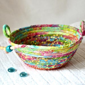 May include: A colorful, handmade fabric bowl with a round shape and two small handles. The bowl is made from strips of fabric in various colors, including pink, green, yellow, and blue. Two small, blue glass stones are next to the bowl.