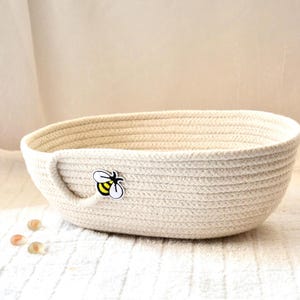 May include: A cream-colored, oval-shaped woven basket with a small handle on one side. A yellow and black bee applique is attached to the side. The basket is made of thick, braided rope and sits on a white surface.