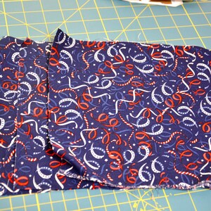 May include: Folded navy blue fabric with a pattern of white, red, and blue ribbons and stars. The fabric is likely cotton or a similar material, and is folded neatly. The fabric is on a green cutting mat.