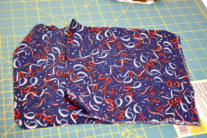 May include: Folded navy blue fabric with a pattern of white, red, and blue ribbons and stars. The fabric is likely cotton or a similar material, and is folded neatly. The fabric is on a green cutting mat.