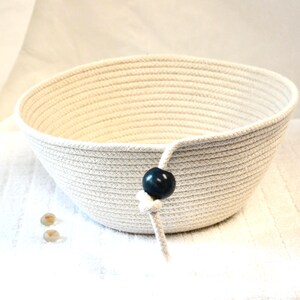 May include: A cream-colored, woven rope bowl with a dark blue bead accent. The bowl has a rounded shape and a textured surface. The bead is attached to the bowl with a white rope. Two small, round, tan-colored stones are in front of the bowl.