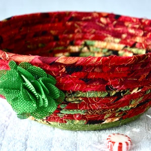 May include: A handmade fabric bowl with a red and floral pattern. The bowl is adorned with a green fabric flower on the side. The bowl is sitting on a white surface. Two wrapped candies are next to the bowl.