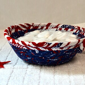 May include: A round, fabric bowl with a patriotic red, white, and blue design, filled with individually wrapped white items. A red star is placed near the bowl. The bowl is on a white, textured surface.