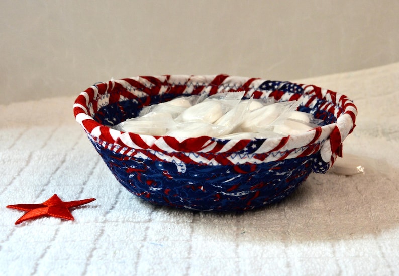May include: A round, fabric bowl with a patriotic red, white, and blue design, filled with individually wrapped white items. A red star is placed near the bowl. The bowl is on a white, textured surface.