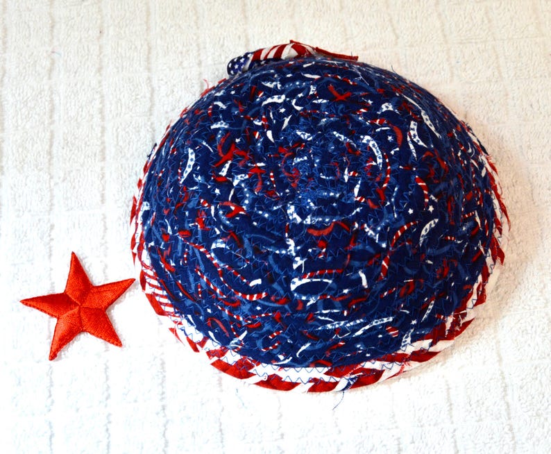 May include: A round, quilted bowl with a patriotic theme. The bowl features a blue fabric with red and white accents, and a red star. The rim is trimmed with red and white striped fabric.