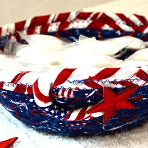 May include: A patriotic fabric bowl with a red star applique. The bowl features a design of red and white stripes, blue fabric with white stars, and is filled with white items. The bowl is likely handmade.
