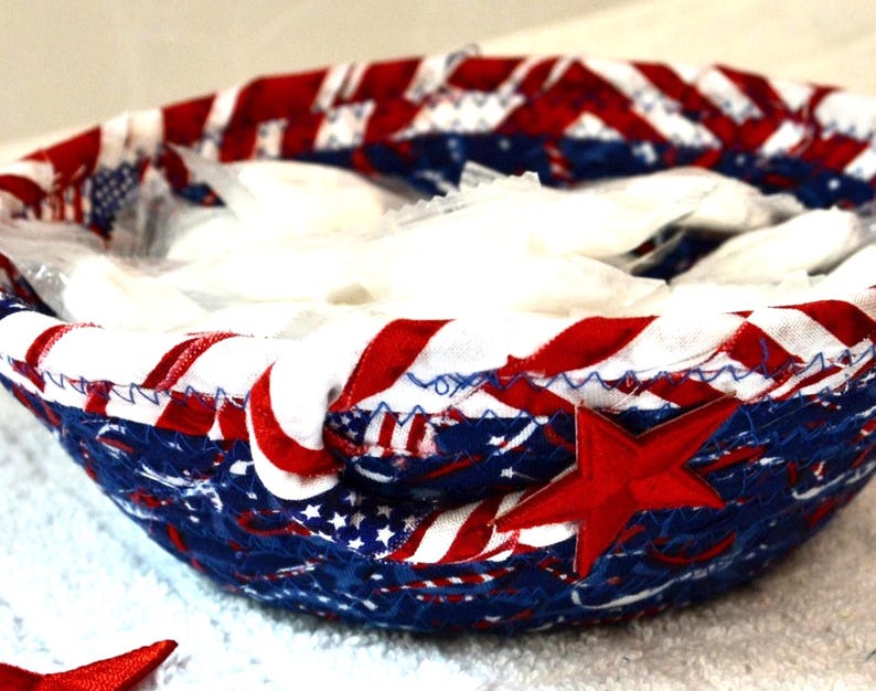 May include: A patriotic fabric bowl with a red star applique. The bowl features a design of red and white stripes, blue fabric with white stars, and is filled with white items. The bowl is likely handmade.