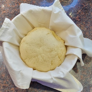 May include: A white cloth-lined bowl with a large, round, pale yellow dough ball inside.
