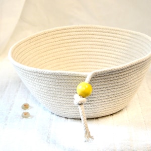 May include: A cream-colored, woven rope bowl with a rounded shape. A yellow bead is attached to the side with a knotted rope detail. Two small, round, light-colored objects are near the bowl. The bowl is likely for home decor or storage.