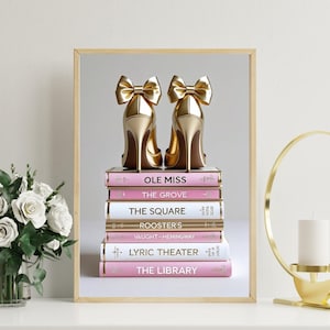 May include: A framed print featuring a stack of books with gold high heels on top. The books are pink and white with gold lettering. The heels are gold with bow accents. The books' titles include "OLE MISS" and "THE GROVE".