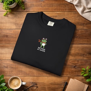 May include: Black crew neck sweatshirt featuring an embroidered cartoon frog wearing goggles and a red scarf. The text "WE RIDE AT DAWN" is below the frog. The sweatshirt is displayed on a wooden surface.