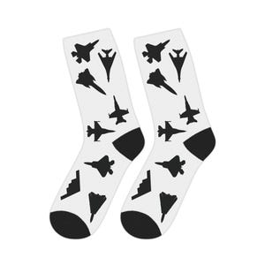 Puede incluir: Un par de calcetines blancos con talones y punteras negras. Los calcetines están estampados con siluetas negras de diferentes aviones de combate. El diseño se repite en ambos calcetines.