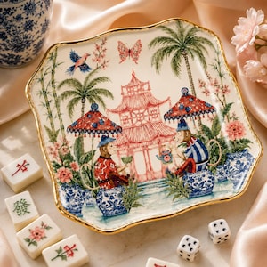 May include: A square ceramic plate with a gold-trimmed scalloped edge, featuring a colourful illustration of two monkeys sitting on blue and white patterned stools under umbrellas, with a pagoda, palm trees, flowers, and a butterfly.