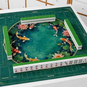 May include: A square green mahjong mat featuring a central circular illustration of koi fish and lotus flowers. Four rows of white and green mahjong tiles are arranged on the mat, with the front row displaying Chinese characters and red symbols.