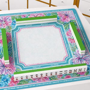 May include: A square mahjong mat featuring a vibrant floral pattern with pink and blue flowers on a teal border. The centre is white with a decorative pink frame. Four rows of mahjong tiles are arranged on the mat, with one row displaying Chinese characters and red and black symbols.