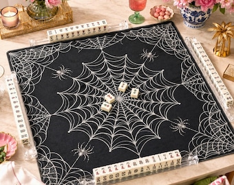 May include: A square black mahjong mat featuring a white spiderweb pattern with several spiders, surrounded by four clear acrylic tile racks holding white mahjong tiles, with a few loose tiles placed in the center of the mat.